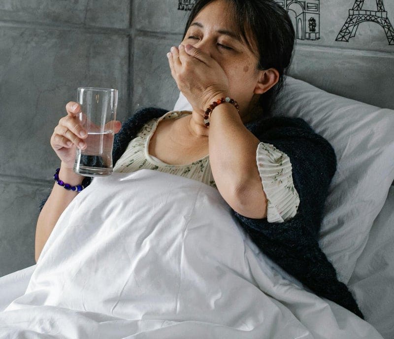 Mujer enferma en la cama, tosiendo, con un vaso de agua en la mano